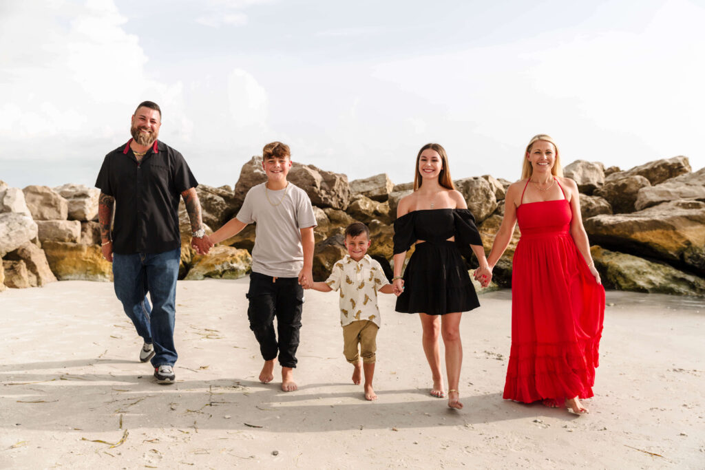 Five people, including two adults and three children, walk barefoot on a sandy beach with rocks in the background, smiling at the camera and holding hands—much like a family enjoying their newly finished garage floor coating together. - Floor Coating Specialist - Bradenton, FL