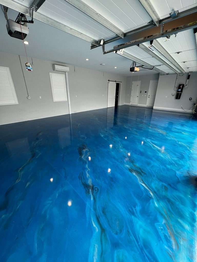 A spacious garage with a glossy, blue epoxy floor coating resembling water, white walls, and an open garage door letting in natural light. - Floor Coating Specialist - Bradenton, FL