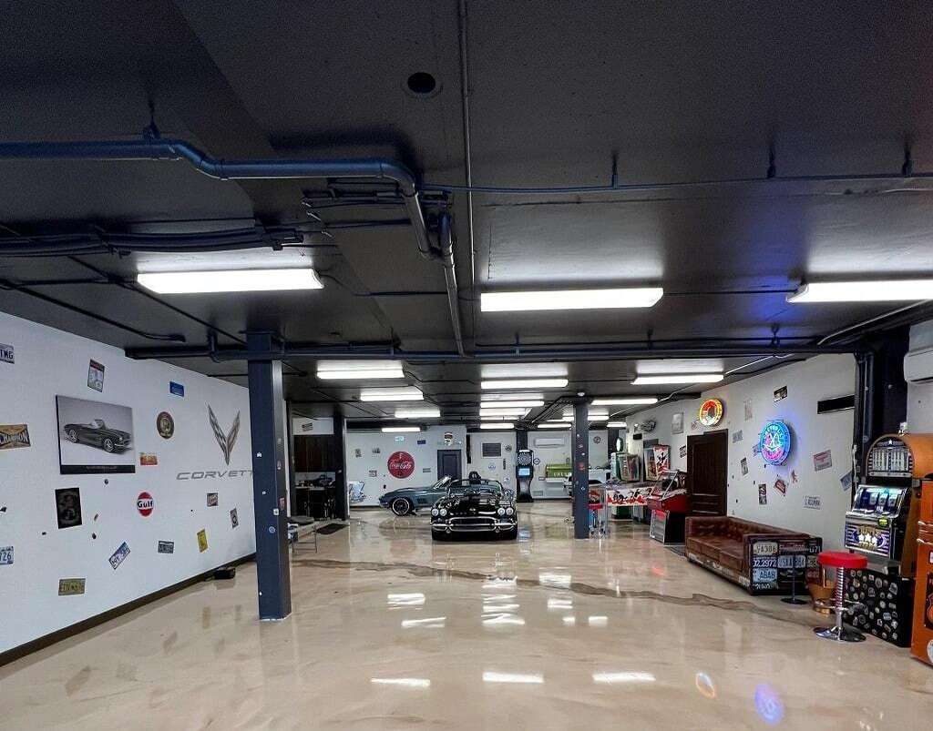 A spacious garage with polished concrete flooring, vintage decor, wall signs, a Corvette logo, classic cars, sofas, and a vending machine. - Floor Coating Specialist - Bradenton, FL