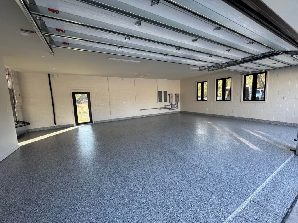 A clean, empty garage with a speckled epoxy floor coating, white walls, multiple windows, garage doors open, and electrical panels on one wall. - Floor Coating Specialist - Bradenton, FL