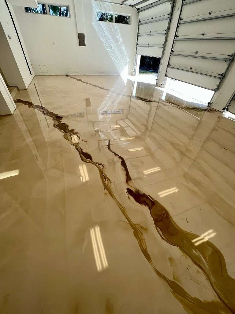 A garage with a glossy, marbled epoxy floor coating featuring brown veining. The space is well-lit, and the garage door is partially open. - Floor Coating Specialist - Bradenton, FL