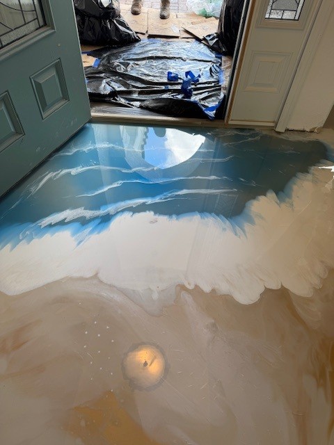 A newly painted entryway floor features a beach and ocean wave design, created with an epoxy floor coating. Painting supplies are visible outside the open door, showcasing the transformation. - Floor Coating Specialist - Bradenton, FL