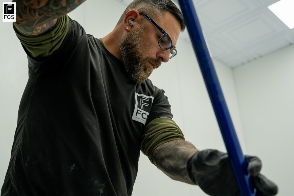 A man with tattoos and a beard, wearing glasses and gloves, works with a blue rod in a bright, white room. The FCS logo is visible on his shirt and in the corner as he applies polyaspartic floor coating to the surface. - Floor Coating Specialist - Bradenton, FL