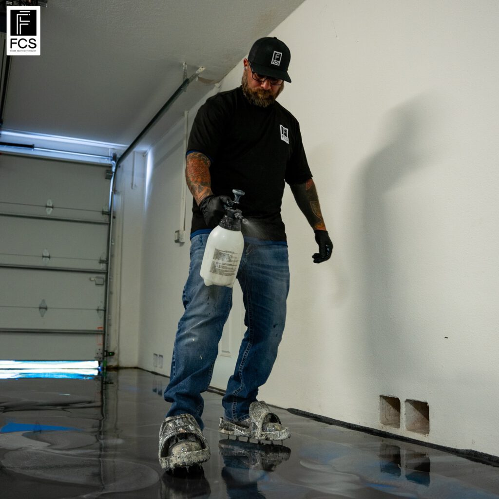 A man wearing spiked shoes and gloves uses a spray bottle to treat an epoxy floor coating, ensuring the shiny garage floor remains spotless and durable. - Floor Coating Specialist - Bradenton, FL