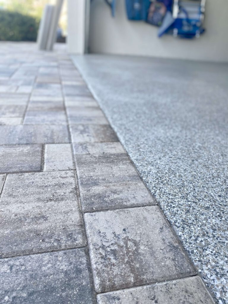 Close-up view of a transition between a textured stone paver driveway and a speckled, finished concrete garage floor with an epoxy floor coating. - Floor Coating Specialist - Bradenton, FL
