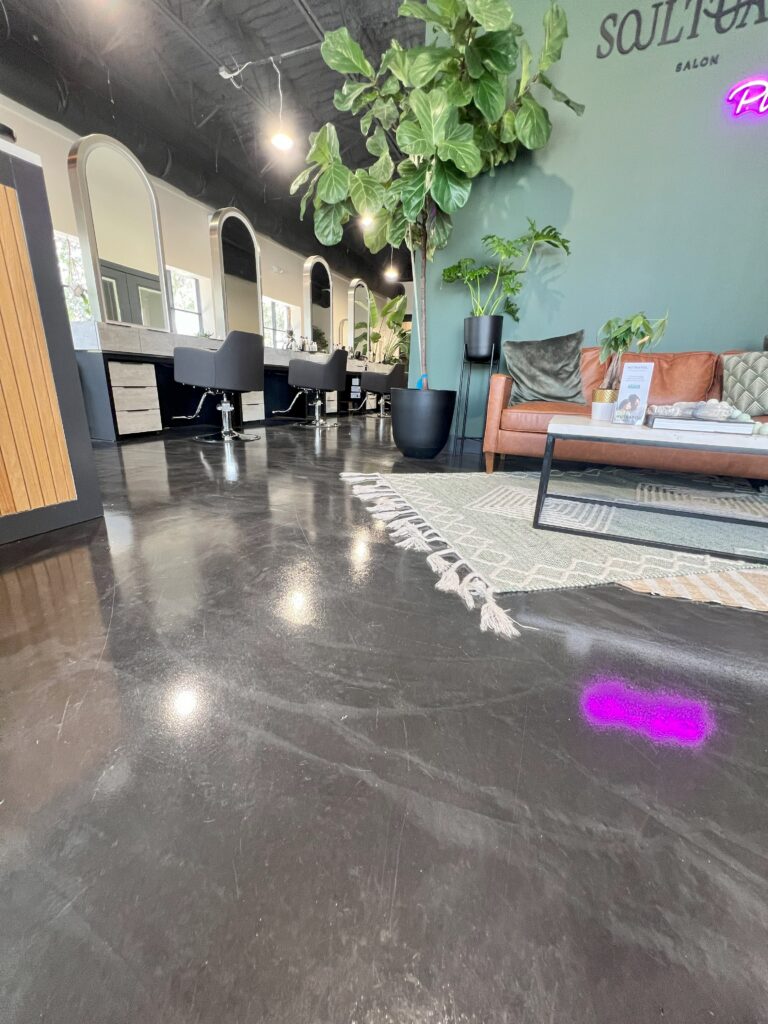 Modern salon interior with black chairs, large mirrors, a potted plant, and a seating area with a couch and rug on sleek polished concrete flooring. - Floor Coating Specialist - Bradenton, FL