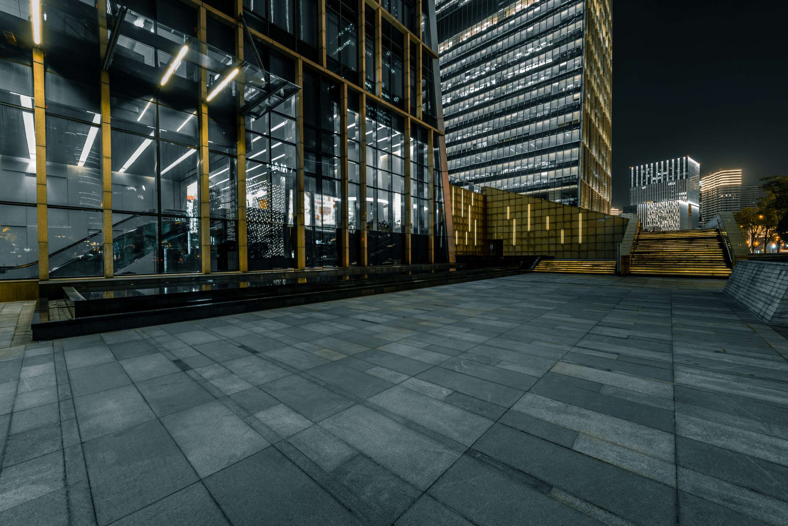 Modern urban plaza at night with illuminated office buildings, glass facades, stone tiles, and steps leading to a higher level. - Floor Coating Specialist - Bradenton, FL