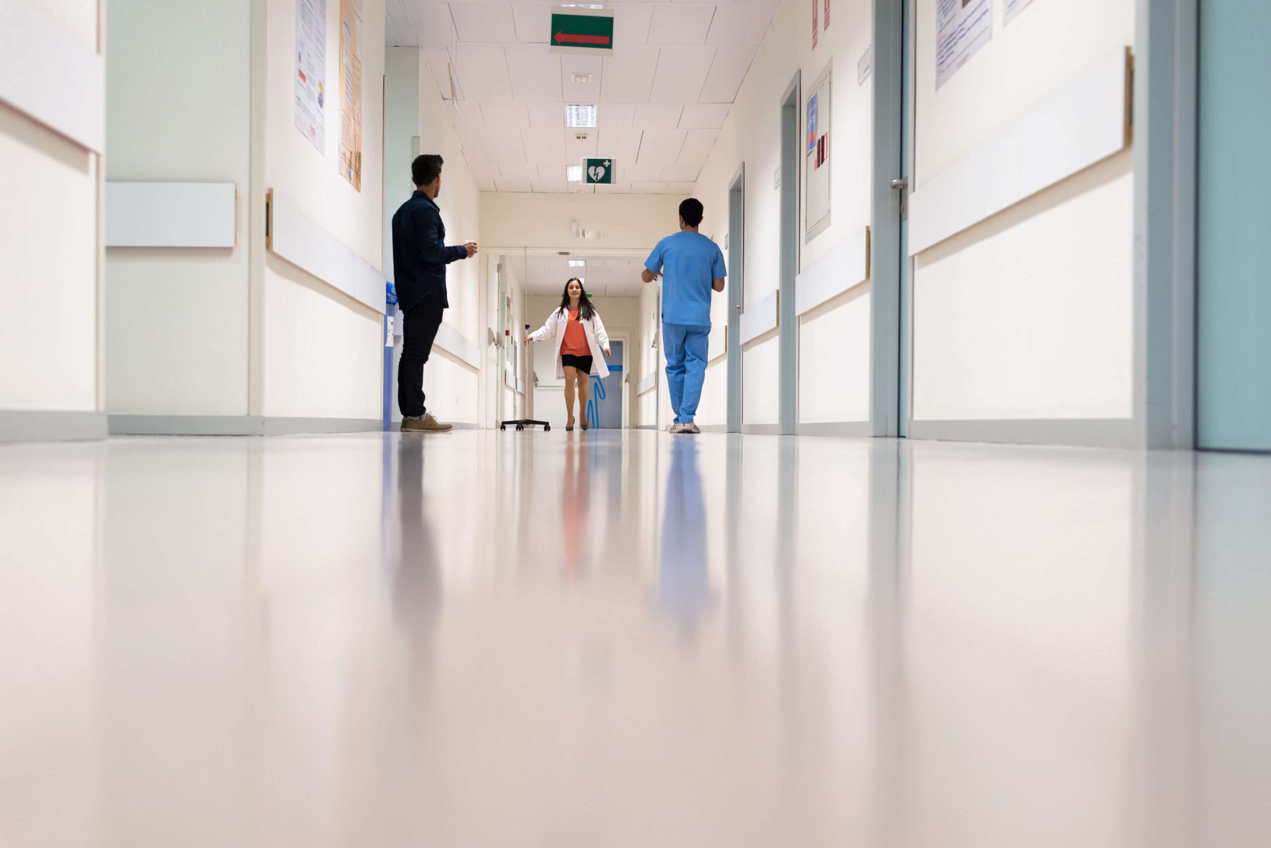 Three people walk in a brightly lit hospital corridor; one wears medical scrubs, and two are in business attire. - Floor Coating Specialist - Bradenton, FL