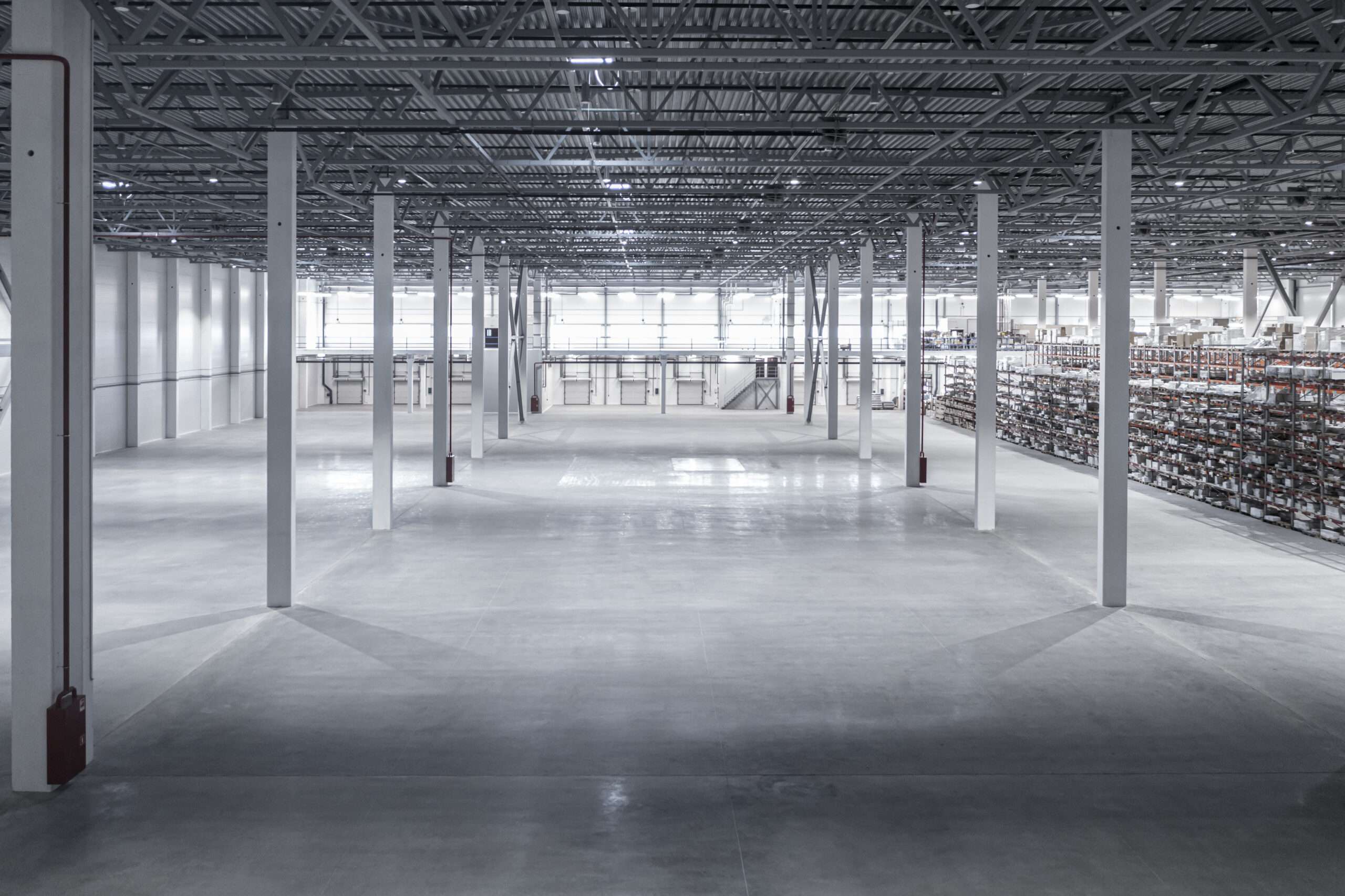 Large empty warehouse interior with concrete floors, white support columns, and visible shelving units along the right wall. - Floor Coating Specialist - Bradenton, FL