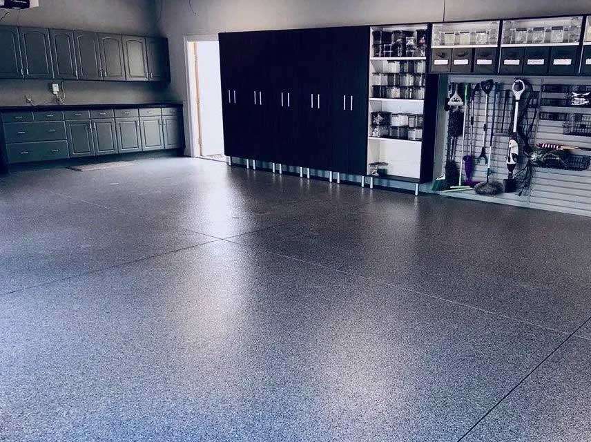 A clean, organized garage with gray cabinets, storage shelves, and a smooth, speckled epoxy floor. Various tools and supplies are neatly arranged along the right wall. - Floor Coating Specialist - Bradenton, FL