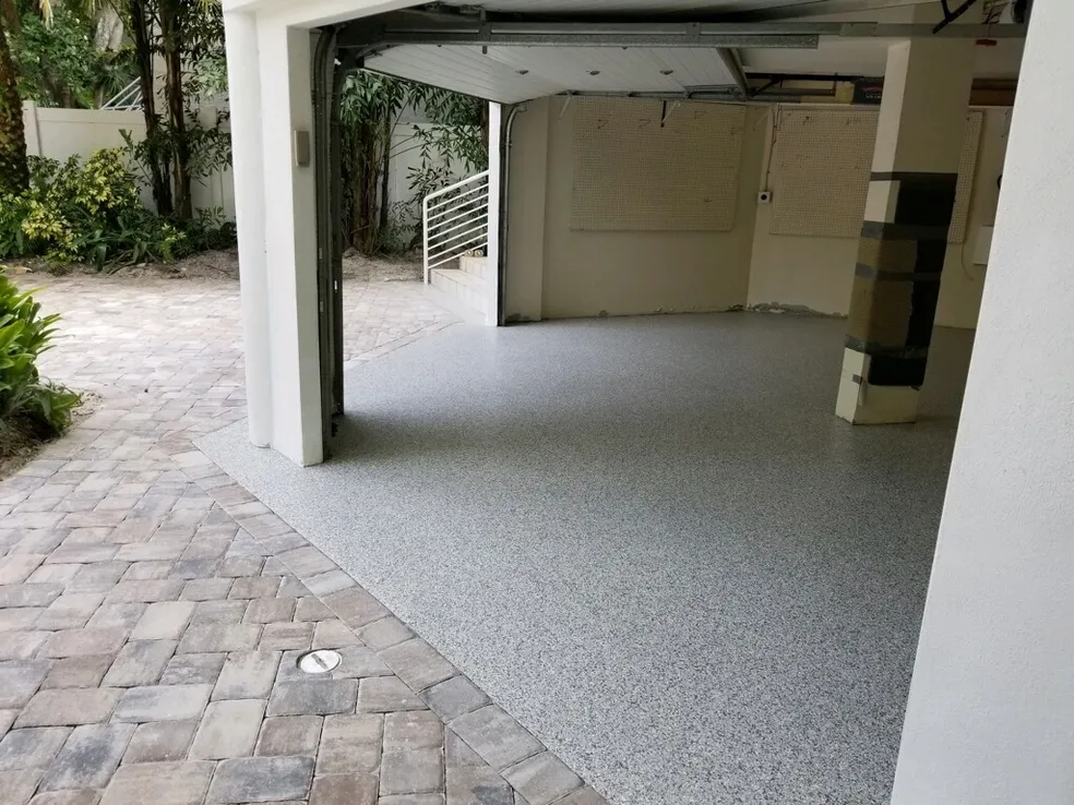 A garage with an open door reveals a clean, speckled epoxy floor coating. The driveway features interlocking stone pavers, and greenery is visible outside. - Floor Coating Specialist - Bradenton, FL