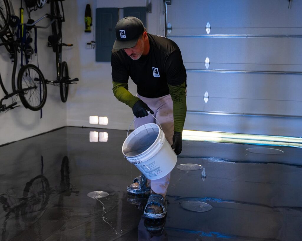 A man wearing protective gear pours liquid from a white bucket onto a shiny, black concrete floor overlay inside a garage, with bicycles and tools visible in the background. - Floor Coating Specialist - Bradenton, FL