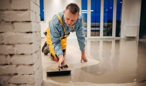 Person in yellow overalls kneeling on the floor, using a tool to spread and smooth wet concrete in a room with large windows. - Floor Coating Specialist - Bradenton, FL