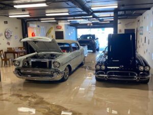 Two classic cars are parked indoors; the car on the left has its hood open, while the black car on the right has its hood raised vertically. Another car is visible in the background. - Floor Coating Specialist - Bradenton, FL