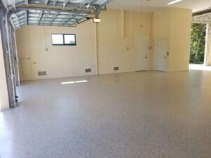 Empty garage with a speckled epoxy floor, cream-colored walls, two white doors, a small window, and an open garage door letting in daylight. - Floor Coating Specialist - Bradenton, FL