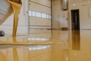 Yellow liquid epoxy is being poured onto the floor of an industrial garage or warehouse, partially covering the concrete surface. - Floor Coating Specialist - Bradenton, FL