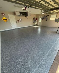 Clean, empty garage with a speckled epoxy floor, wall-mounted storage cabinets, a hanging bicycle, and various items on the walls. - Floor Coating Specialist - Bradenton, FL