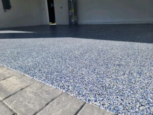 Close-up view of a garage floor coated with a blue and gray speckled epoxy finish, next to a row of gray pavers at the entrance. - Floor Coating Specialist - Bradenton, FL